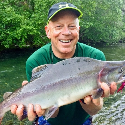 Todd smiling and holding a fish in hand infront of a river and green trees 