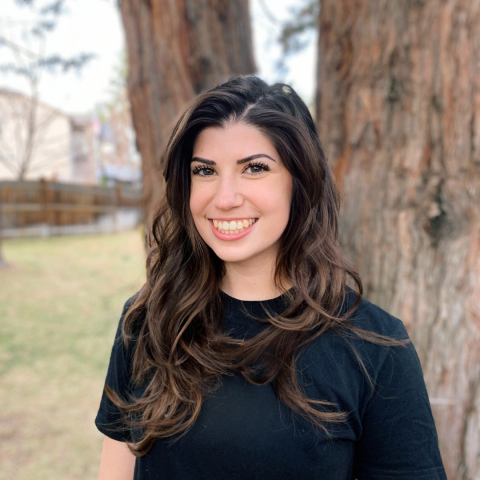 A woman with dark hair smiles and looks toward the camera. She is outside standing in front of a tree trunk.