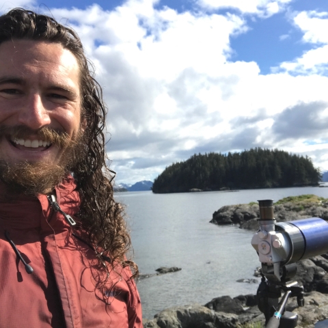 A man standing next to a spotting scope with a rocky shoreline and island in the background