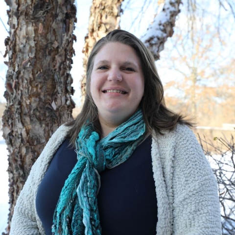 a woman wearing a scarf and sweater smiles and stands outside