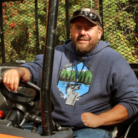 A bearded, smiling man in a sweatshirt with a leaping salmon pictured on it sits in an ORV with a Service ballcap and sunglasses perched on the cap's rim.