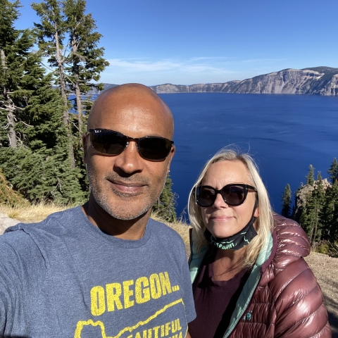 A man and woman smile, both wearing sunglasses, with a deep blue lake, mountains, and trees in the background