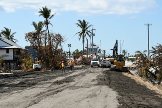 Heavy equipment parked on damaged asphalt alongside construction debris and damaged palm trees.