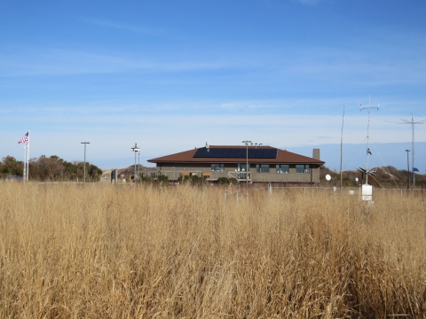 Grassy field with a brown building in the background. There are solar panels on the roof of the building. An American flag is flying on the left side of the image.