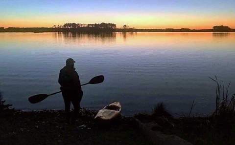 A kayaker on the bank of a marsh with a sunset behind a few stands of pine trees in the distance