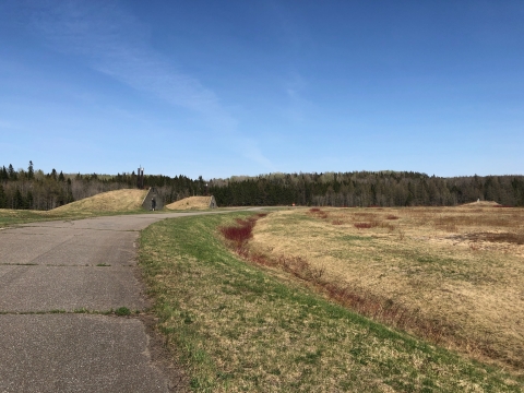 A paved driveway loops past two military bunkers in a mowed field with conifer trees seen distant on the horizon. 