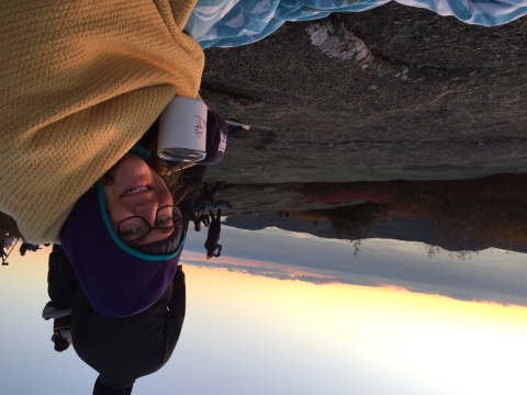 Rachel Stearns, biological technician at Rachel Carson National Wildlife Refuge, savors a cup of coffee as the dawn breaks in the distance. Rachel is huddled against the cold in a bright yellow blanket. 