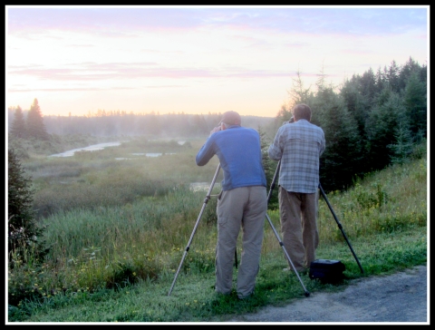 Two men with a wildlife spotting scope and camera are positioned at an overlook at dusk. Within view of is a pond ringed with evergreens. 