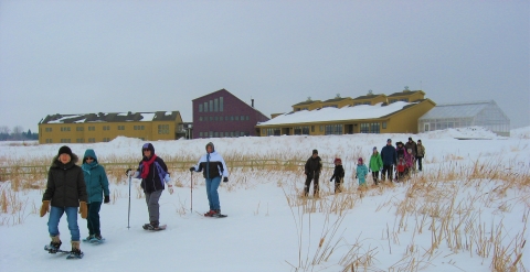 A line of about a dozen children and adults snowshoe across a bridge 