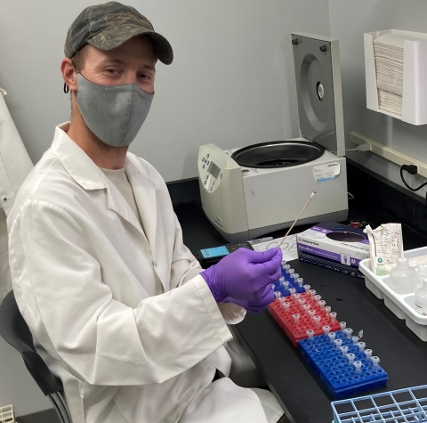 A person in a hat, mask and a tray of tubes at a lab table