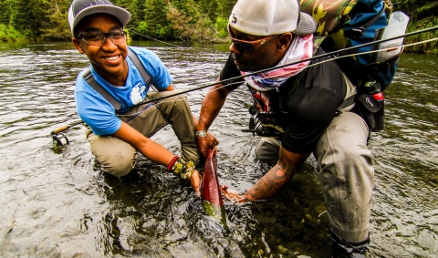 A man and a boy with a fly fishing rod kneel together in a stream, holding a fresh-caught salmon.