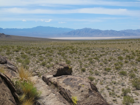 a rock in the foreground serves as a lookout across a desert landscape with a dry lakebed and mountains in the distance