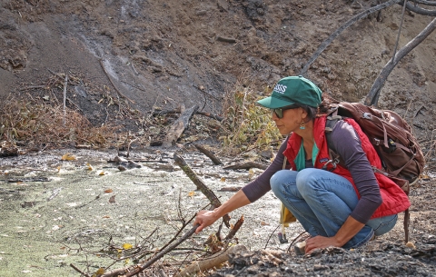 A woman crouches next to a river holding a stick