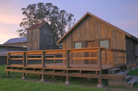 A wooden cabin with the deck in the foreground and a wooden barn-like structure behind it