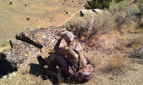 Woman dressed in camouflage sitting on the ground. Looking for animals in the valley below her with binoculars