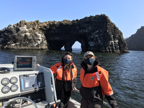 Two young women in orange float coats in a boat in front of a coastal island smile at the camera 