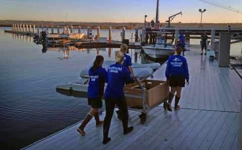 Four uniformed people roll dolly with wooden crate down a dock