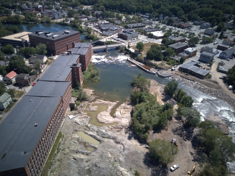 Aerial view of a river flowing through a city. Waterfalls, and a fishway channel are visible. Red brick buildings line the river bank. 