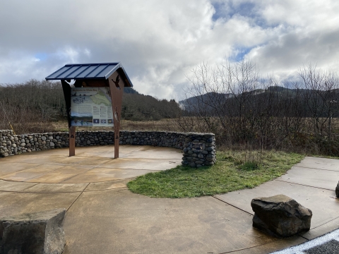 A kiosk with a background of clouded blue sky over forested hills and grassy tidelands