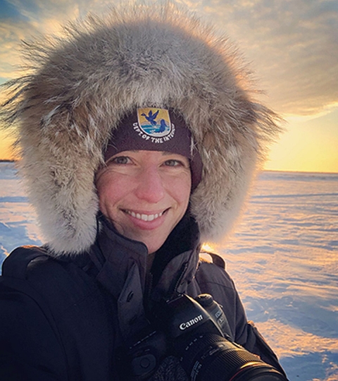 Close-up of a woman standing on an arctic snow field. She bundled up for winter and has a camera handing from her neck