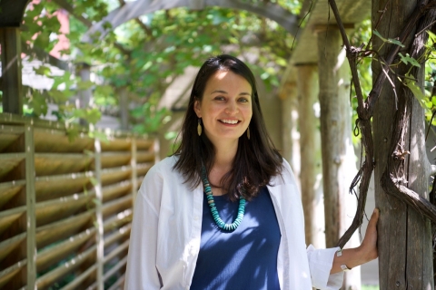 Smiling, Bonney Hartley faces the camera. She has dark hair and wears blue and white. She stands in partial shade underneath a rounded wooden trellis.