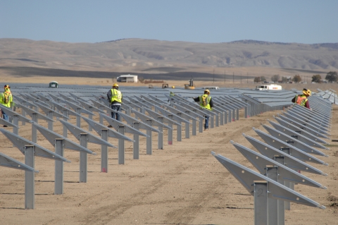 rows of gray solar panels stand out against the dry desert background. Solar panel installers wearing yellow vests work on the panels.