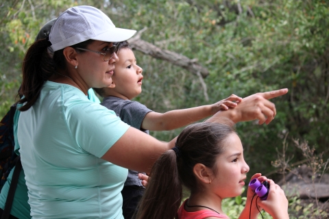 A woman wearing a sun visor and accompanied by her young daughter and younger son points to birds they see off camera.s