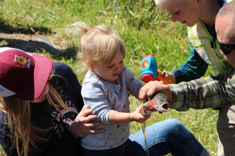 A family surrounds a toddler grasping a rainbow trout her father holds for her. Mom steadies her, and brother hands her a plastic toy hammer.