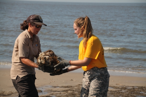 This photo shows two women standing on a beach. One (image left) hands a mesh bag filled with shells to the other (image right). The ocean lapping against the beach is visible behind them. 