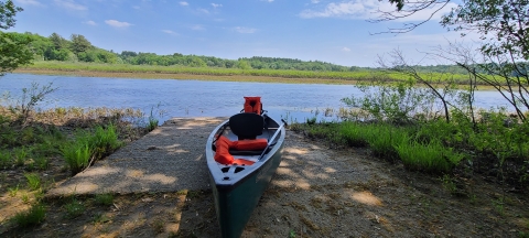 Photo of the canoe launch off of Weir Hill Trail, with a dark blue canoe sitting on the ground in front of the Sudbury River 