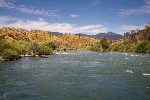 a landscape of a river with hills in the background udner a cloudy blue sky