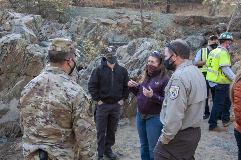 Several people wearing masks talk outside, including one in camouflage