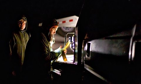 Two men read a document by a small light in the dark