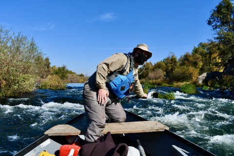 A man wears a mask while standing at the front of a boat on a swift moving river