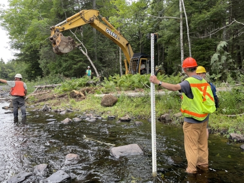 biologist standing in river with excavator in the background