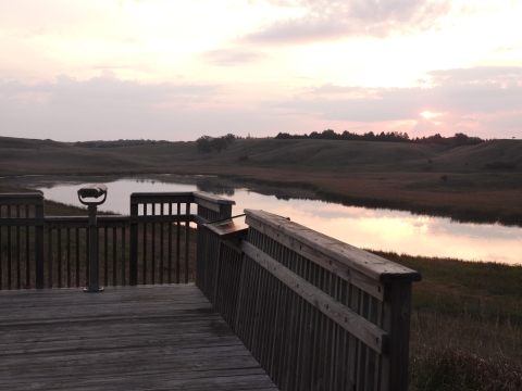 A wooden observation deck with a spotting scope overlooks a wetland at the base of some rolling hills