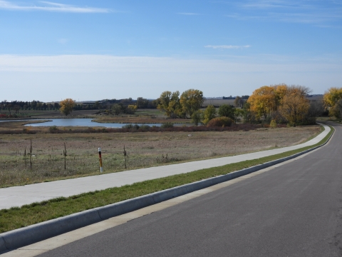 A paved path next to the road follows the edge of the prairie and disappears in the distance amongst a group of trees. A small lake can be seen in the distance.