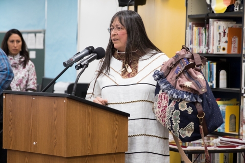 Woman wearing traditional white garment stands at podium holding small cloth bag.