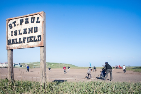 Sign in the foreground reads St. Paul Island Ballfield and people are on a dirt playing field with a blue sky overhead.