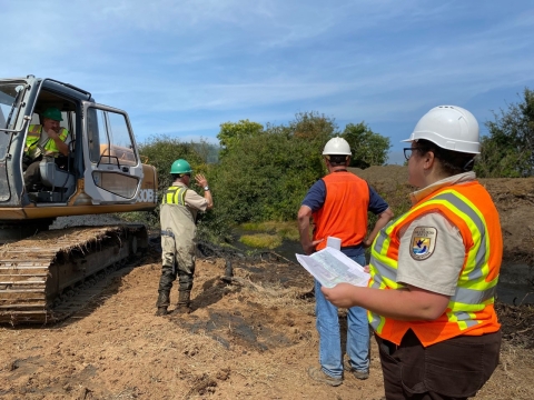 Men in helmets and reflective vests stand near a piece of heavy equipment and look at plans.