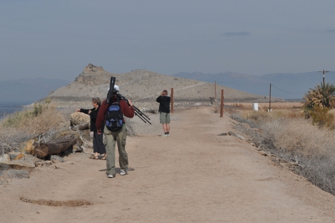 A person carries a spotting scope on a tripod along a trail toward two other birdwatchers with binoculars. There is open water to the left of the trail and a large, rocky hill in the distance. 