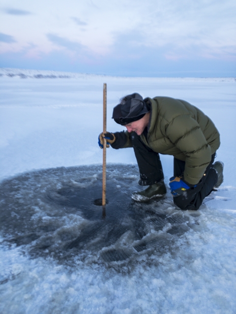 a man crouches near a small round hole in the ice and uses a yardstick to measure 