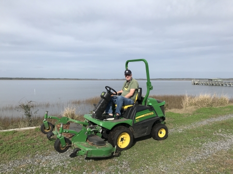 A man sits on a large green lawn mower with water and a dock in the background.