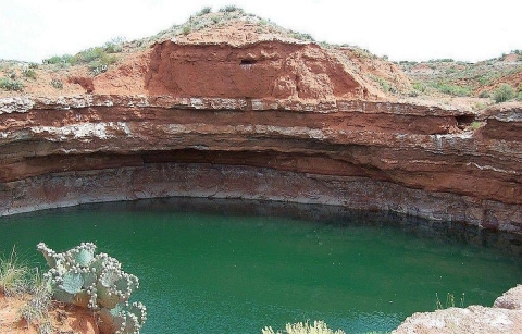 A large sinkhole full of aqua-green-colored water in a rocky, hilly desert landscape