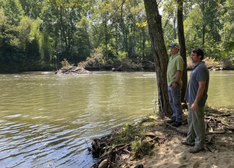 Two men standing in the shade of trees on river bank admiring peaceful green landscape.