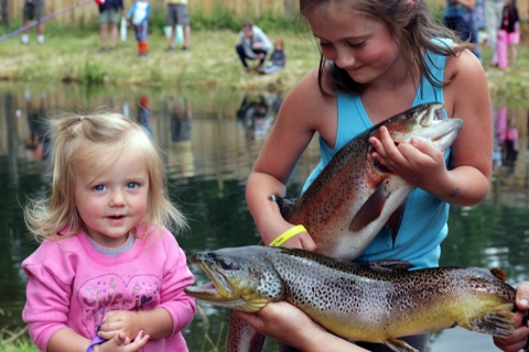 Two girls pose with two big trout in front of a pond where other children are fishing.