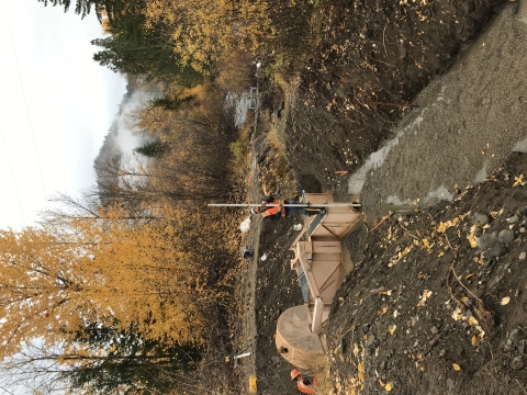 A man in orange vest and hardhat stands on a structure in a dewatered streambed with a tall white rod.