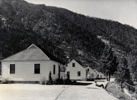 Black and white photo of 4 small, wood-sided, white-painted houses on a low hill, surrounded by closely-cropped lawn. A man reclined on his elbows at the edge of the lawn, facing the view.