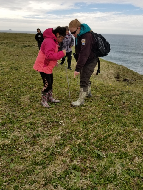 Woman in brown USFWS uniform instructs child wearing bright pink coat with water and sky in the background