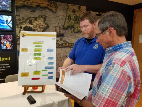 Two men flip through pages as they stand in front of a table. One is wearing a uniform.